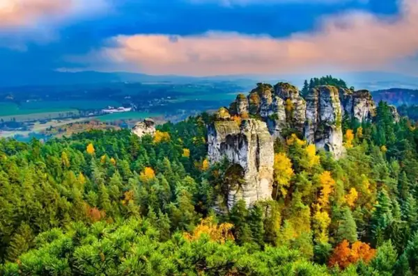 Felsformationen im böhmischen Paradies (Český ráj) ragen aus einem bunten Herbstwald hervor, unter einem dramatischen Himmel.