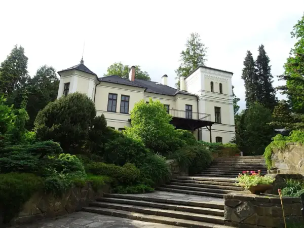 Ein hellgelbes Herrenhaus mit Turm und Balkon, umgeben von üppigem Grün und einer Steintreppe, in Opava, Tschechien.