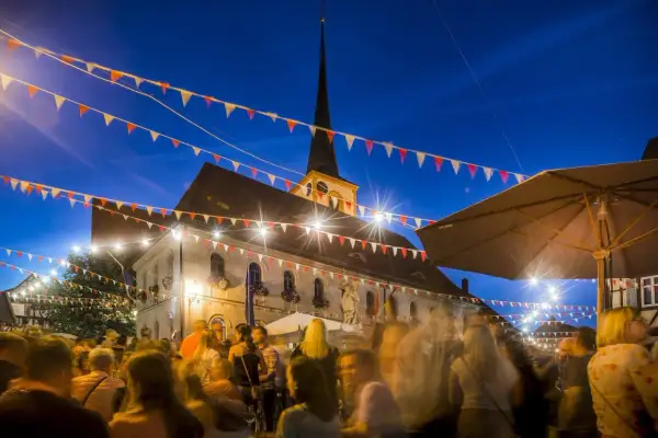Bunte Girlanden schmücken den Abend bei Weinfeste Franken. Menschen feiern vor einem beleuchteten Gebäude mit Kirchturm.